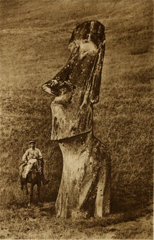 Photograph of a moai statue standing on the grassy slope of Rano Raraku, Easter Island, with a boy on horseback beside it for scale